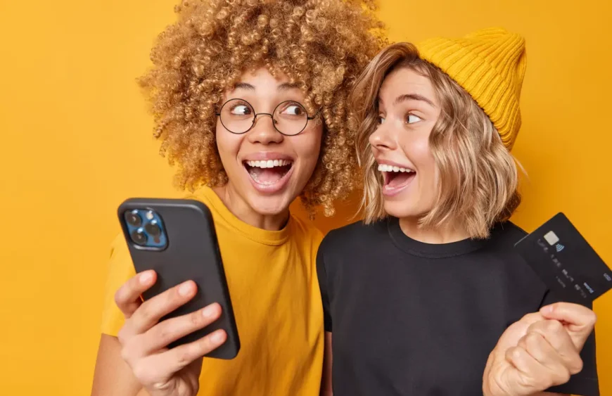 positive-glad-female-friends-holds-credit-card-smartphone-happy-spend-money-online-shopping-purchase-goods-services-internet-isolated-yellow-background-check-their-balance (1)